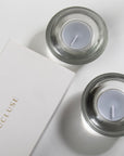 An overhead top-down view of two grey blue velvet tealight candles resting in modern clear glass holders next to a white embossed Vaucluse gift box.