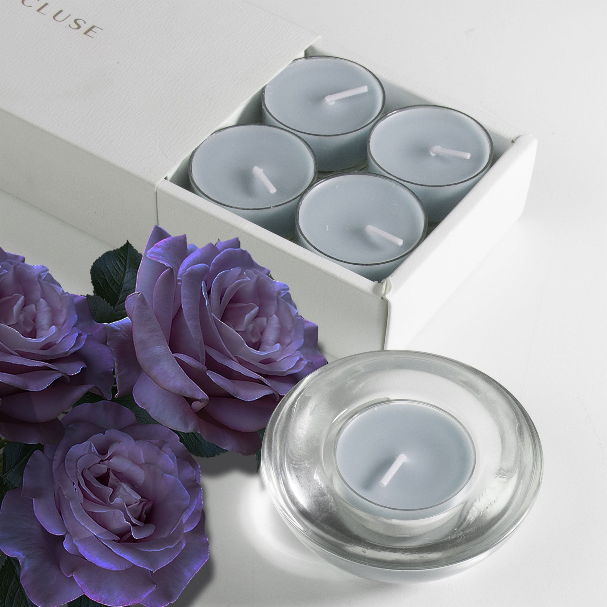 A grey blue velvet scented tealight candle in a clear glass holder in the foreground, styled with deep purple roses next to the sliding tray packaging.