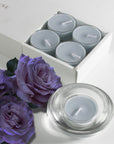 A grey blue velvet scented tealight candle in a clear glass holder in the foreground, styled with deep purple roses next to the sliding tray packaging.