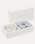 An open luxury white gift box displaying a set of four soft grey velvet tealight candles next to a round clear glass candle holder.