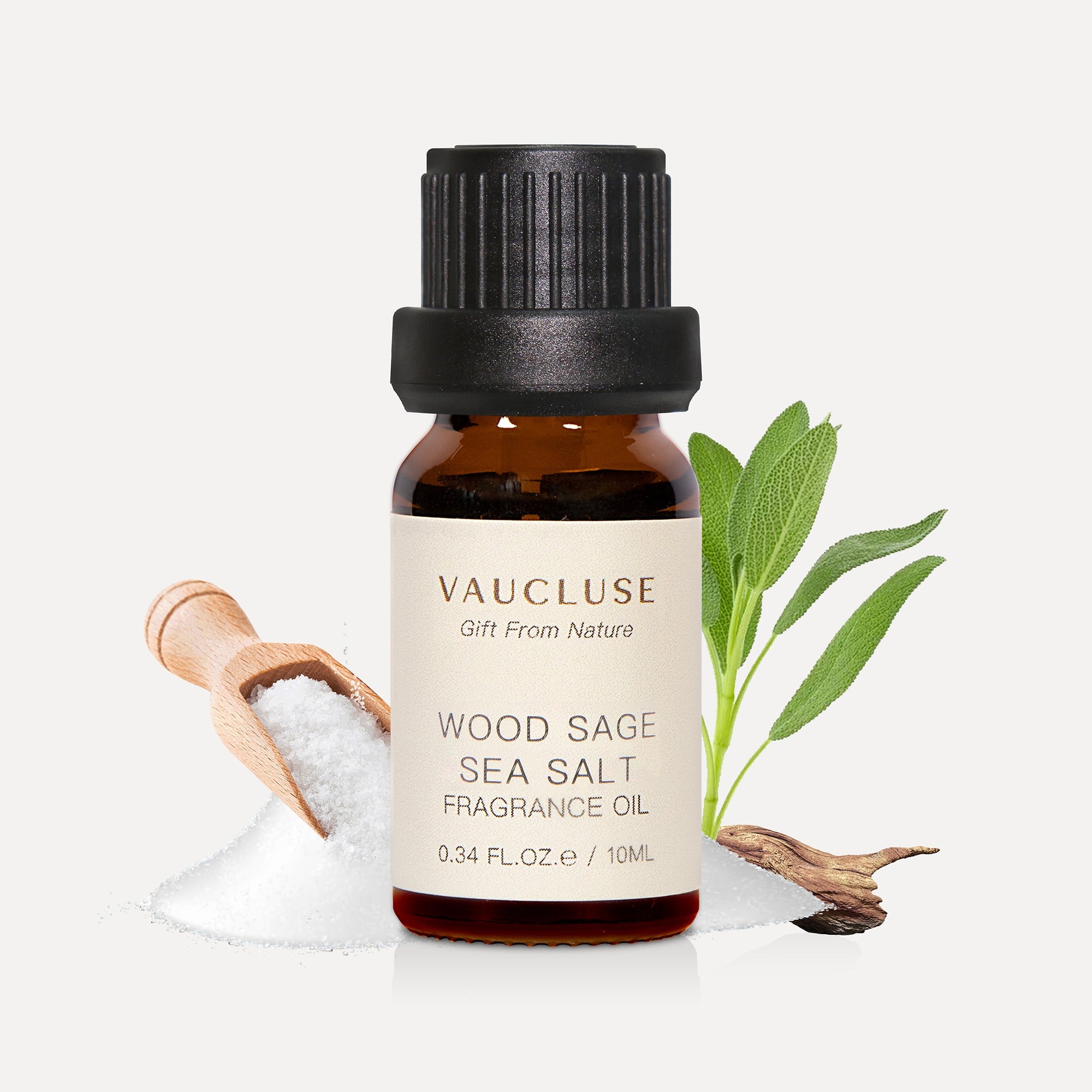 A 10ml amber glass bottle of Vaucluse Wood Sage Sea Salt Essential Oil backed by green sage leaves and a wooden scoop filled with coarse sea salt.