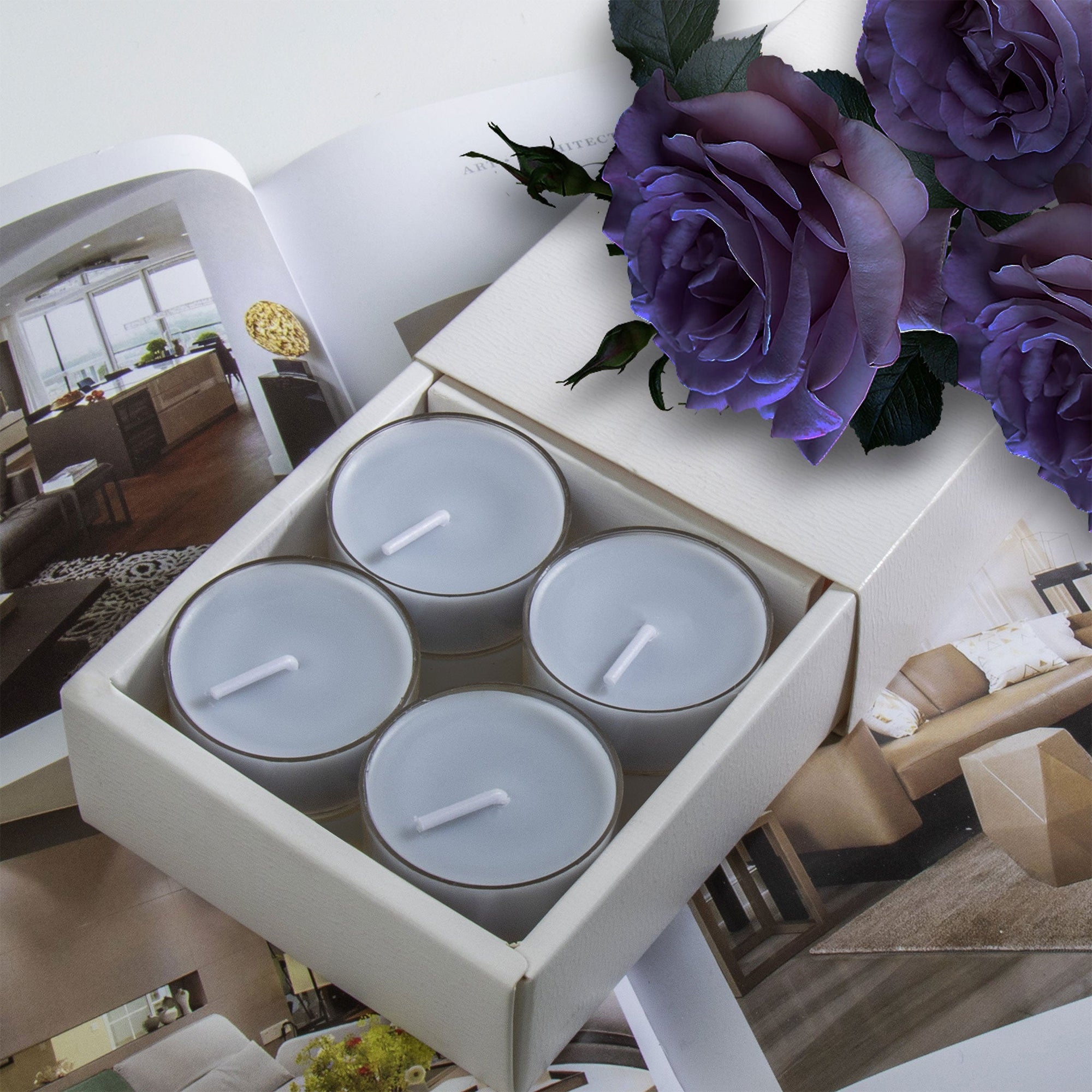 A premium white Vaucluse 16-pack box sliding open to reveal a tier of grey blue velvet tealight candles, elegantly styled beneath soft purple roses.