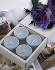 A premium white Vaucluse 16-pack box sliding open to reveal a tier of grey blue velvet tealight candles, elegantly styled beneath soft purple roses.