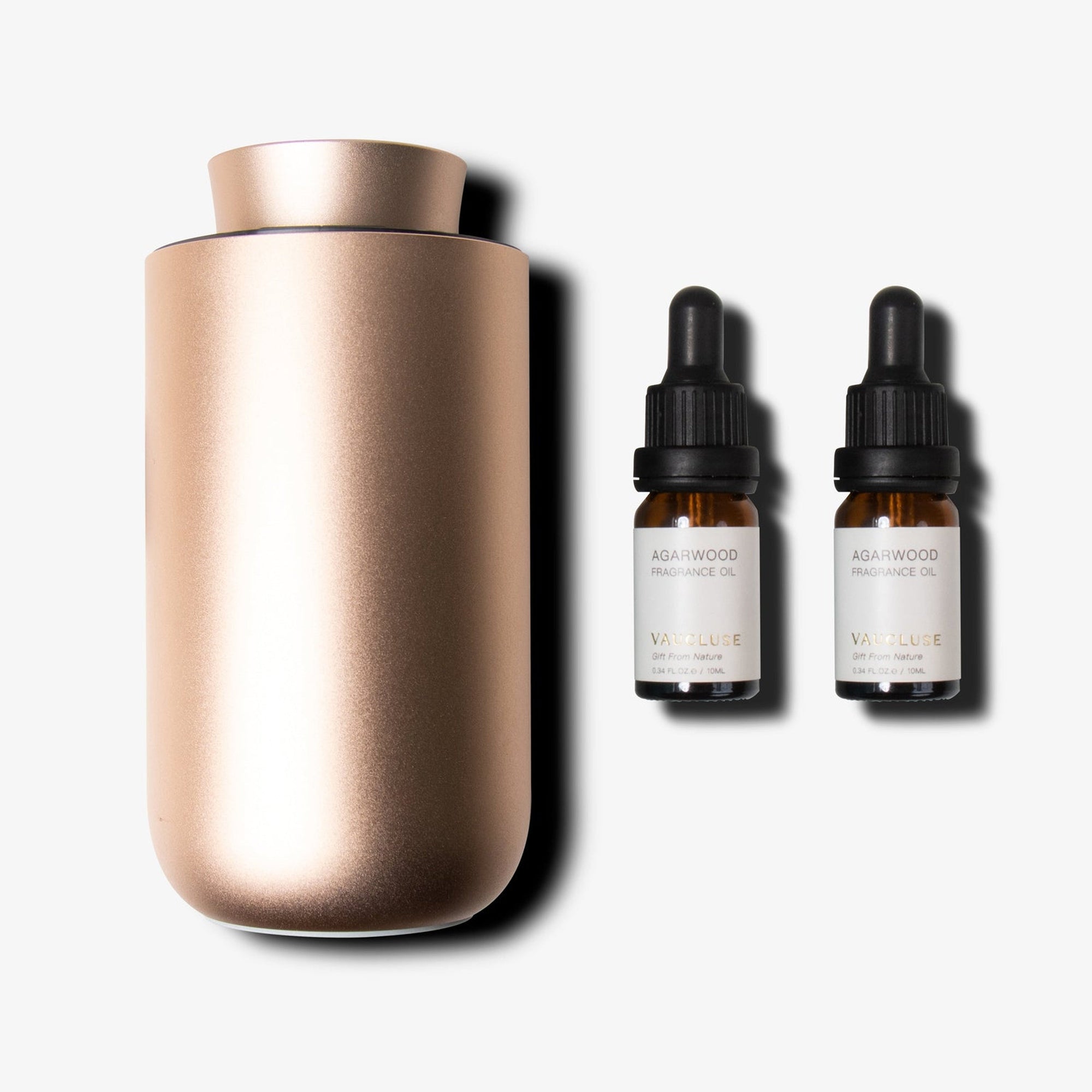 A premium rose gold waterless diffuser shown next to two small amber glass essential oil dropper bottles with clean cream color labels.